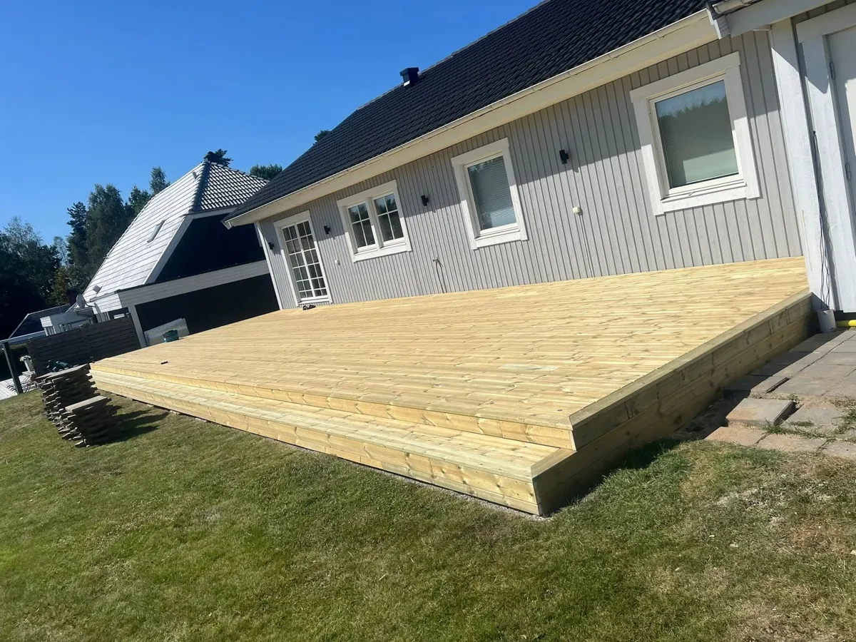 Ny trall i tryckimpregnerat trä längs en ljusgrå villa en solig dag