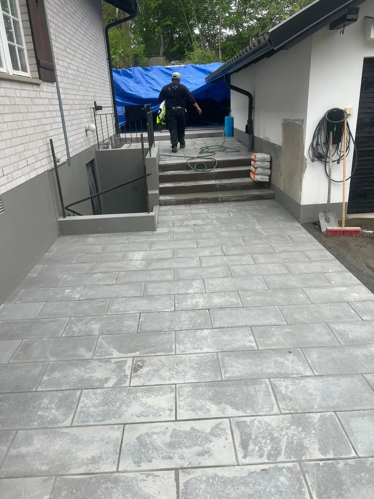 Plattsatt yta framför en trappa, där en hantverkare går upp mot huset